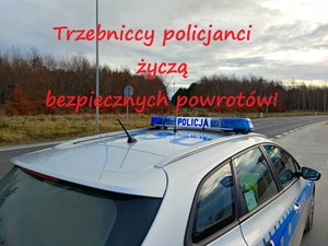 Oznakowany radiowóz policyjny stoi przy drodze. Nad nim znajduje się napis Trzebniccy policjanci życzą bezpiecznych powrotów.