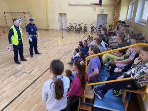 Policjanci stoją przed dziećmi i omawiają teorię. Dzieci siedzą na ławkach przy ścianie. Spotkanie odbywa się na sali gimnastycznej