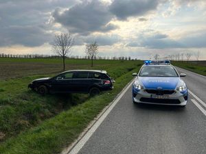 Zdjęcie przedstawia radiowóz oznakowany stojący na jedni, a obok poza jednią stoi auto biorące udział z kolizji. Pojazd jest uszkodzony.