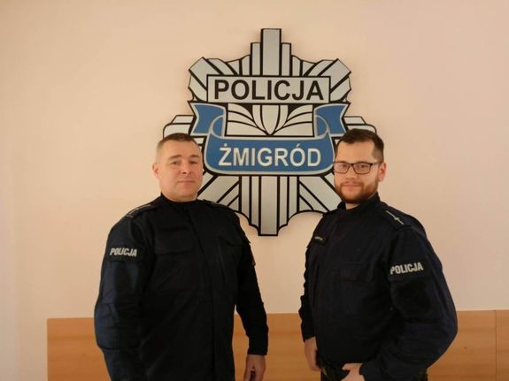 Zdjęcie przedstawia dwóch policjantów wyróżnionych przez Komendanta Wojewódzkiego Policji we Wrocławiu. Policjanci ubrani są w mundury granatowe ćwiczebne. Stoją w pomieszczeniu a między nimi na ścianie wisi gwiazda policyjna z napisem Policja Żmigród