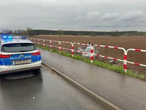 Zdjęcie przedstawia radiowóz oznakowany stojący tyłem do zdjęcia z włączonymi światłami. Z prawej strony w polu znajduje się samochód osobowy, który jest uszkodzony.