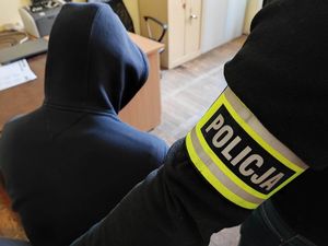 Mężczyzna siedzi na krzesełku tyłem do zdjęcia. Na głowie ma założony kaptur. obok niego stoi policjant który na ręce ma założoną opaskę odblaskową z napisem Policja