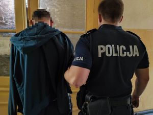 Poszukiwany mężczyzna stoi tyłem do zdjęcia ma założoną bluzę oraz niewidoczną głowę. Obok niego stoi policjant również tyłem do zdjęcia w mundurze służbowym.