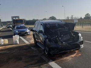 Na drodze ekspresowej, na lewym pasie stoi osobowy mercedes, który ma uszkodzony cały przód auta w wyniku tego, że najechał na tył ciężarówki. Za nim stoi oznakowany radiowóz, za radiowozem pomoc drogowa. Na drodze rozsypany jest proszek oraz ustawione są pojemniki pomocy drogowej.