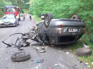 Auto biorące udział w wypadku leży na dachu na drodze. Obok niego znajduje się wiele elementów od samochodu. Przed autem stoi radiowóz oraz pojazd straży pożarnej.