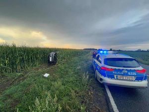 Samochód leży na lewym boku w polu kukurydzy, na jezdni stoi oznakowany radiowóz policyjny z włączonymi światłami błyskowymi.