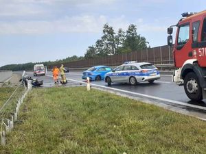 Uszkodzony samochód osobowy stojący na drodze ekspresowej S5,  oznakowany radiowóz policyjny oraz zastęp Straży Pożarnej. W tle stoi karetka pogotowia ratunkowego