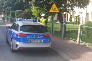 Oznakowany radiowóz policyjny obok niego stoi policjant umundurowany w odblaskowej kamizelce. Policjant sprawdza oznakowanie w rejonie szkoły.