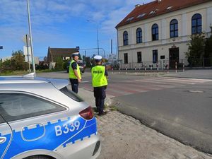 Policjanci w mundurach służbowych oraz kamizelkach odblaskowych stoją w okolicy przejścia dla pieszych. W tle budynek szkoły.