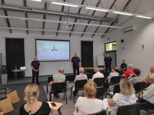 Policjanci stoją przed rzutnikiem w sali biblioteki, przed nimi siedzą zaproszeni goście