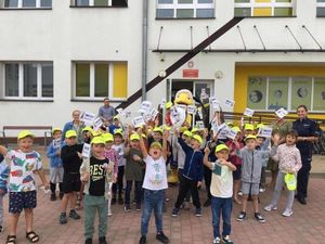 Dzieci stoją przodem do zdjęcia wraz z policjantką oraz maskotką mrówki. Zdjęcie zrobione przed budynkiem szkoły.