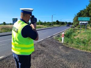 Policjant ruchu drogowego ubrany w mundur oraz kamizelkę odblaskową dokonuje pomiaru prędkości na drodze trzymając w rękach urządzenie do pomiaru prędkości.