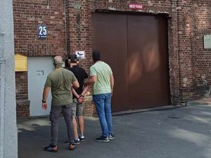 Nieumundurowani policjanci, którzy mają założone kominiarki na twarzy stoją z zatrzymanym poszukiwanym przed wejściem do Aresztu Śledczego.