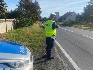 Policjant ruchu drogowego ubrany w mundur służbowy oraz kamizelkę odblaskową dokonuje pomiaru prędkości na drodze dwukierunkowej. Po lewej stronie widoczny przód radiowozu oznakowanego