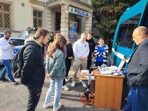 Uczniowie zapoznają się z pracą technika kryminalistyki. Policjant w ubraniu cywilnym stoi wraz z młodzieżą na zewnątrz przed oznakowanym radiowozem. Na stole leży sprzęt wykorzystywany do zabezpieczania śladów.