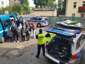 Zdjęcie zrobione z góry na parking Komisariatu Policji w Obornikach Śląskich, gdzie ustawione są radiowozy służbowe. Po lewej stronie stoją uczniowie obok technika kryminalistyki. Po prawej stronie znajdują się policjanci ruchu drogowego przy oznakowanym radiowozie policyjnym. Bagażnik jest otwarty, a w nim znajduje się sprzęt służbowy.