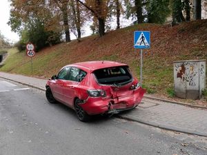 Czerwony pojazd stoi częściowo na chodniku. Tył auta jest uszkodzony, a szyba jest wybita.