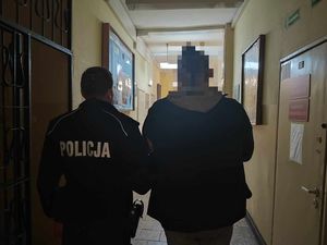 Policjant w mundurze służbowym stoi obok poszukiwanego mężczyzny. Oboje stoją tyłem do zdjęcia. Głowa mężczyzny jest zamazana.