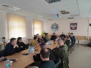 przy stole siedzą i prowadzą konwersację umundurowani funkcjonariusze policji, straży pożarnej, straży leśnej, straży rybackiej, straży ochrony kolei, wojska, nadleśnictwa