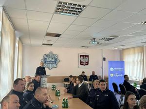 Komendant Powiatowy Policji w Trzebnicy komisarz Grzegorz Maciąg przy mównicy omawia stan bezpieczeństwa w na terenie powiaty trzebnickiego. Przy stołach siedzą policjanci oraz zaproszeni gości.