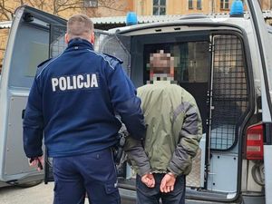 Tyłem do zdjęcia stoi policjant w mundurze służbowym oraz zatrzymany mężczyzna. Stoją na tle oznakowanego radiowozu typu bus.