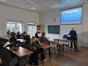 Policjantka ubrana w mundur służbowy chodzi pomiędzy ławkami szkolnymi w których siedzą uczniowie. Na ścianie wyświetlona jest prezentacja multimedialna.