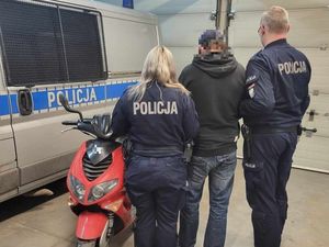 W garażu przy oznakowanym radiowozie policyjnym typu bus tyłem do zdjęcia stoją umundurowani policjanci. Pomiędzy nimi stoi również odzyskany motorower.