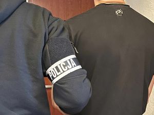 Tyłem do zdjęcia stoi nieumundurowany policjant, który ma na ramieniu opaskę z napisem POLICJA, obok niego stoi zatrzymany mężczyzna. Zdjęcie obejmuje tylko plecy.