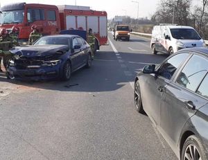 Zdjęcie wykonane na drodze, gdzie miała miejsce kolizja drogowa. Samochód osobowy z uszkodzonym całym przodem, za nim stoi radiowóz Straży Pożarnej oraz funkcjonariusze. Z prawej strony widoczny nieoznakowany radiowóz policyjny oraz przejeżdżające auta.