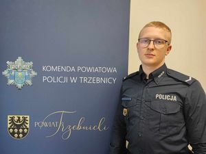 Policjant w mundurze służbowym stoi obok baneru Komendy Powiatowej Policji w Trzebnicy