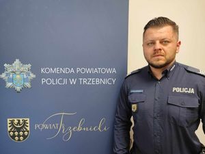 Policjant w mundurze służbowym, stoi obok baneru Komendy Powiatowej Policji w Trzebnicy