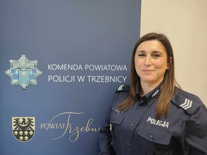 Policjantka w mundurze służbowym stoi przy banerze Komendy Powiatowej Policji w Trzebnicy. Zdjęcie zrobione w pomieszczeniu budynku
