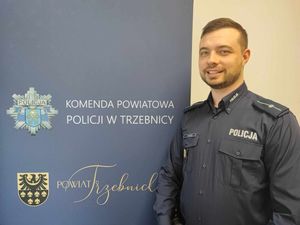 Policjant w mundurze służbowym stoi obok baneru Komendy Powiatowej Policji w Trzebnicy