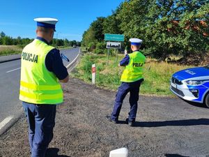 Dwóch policjantów ubranych w mundury służbowe oraz kamizelki odblaskowe stoi na poboczu jedni. Ustawieni są tyłem do zdjęcia.