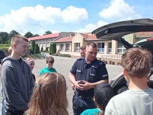 Na placu przed budynkiem szkoły policjant stoi obok radiowozu i rozmawia z dziećmi