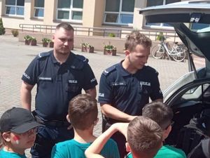 Na placu przed budynkiem szkoły policjant stoi obok radiowozu i rozmawia z dziećmi