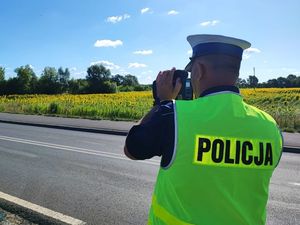 Policjant ruchu drogowego ubrany w kamizelkę odblaskową w ręce trzyma urządzenie do pomiaru prędkości. Policjant stoi przy drodze, a w tle widoczne jest pole zakwitniętego rzepaku