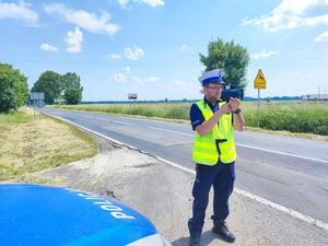 Policjant ruchu drogowego dokonuje pomiaru prędkości. W rękach trzyma urządzenie do pomiaru.. Pogoda słoneczna w tle pola. Policjant stoi przy drodze