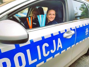 Dwie policjantki siedzą w oznakowanym radiowozie, w którym jest otwarta przednia szyba. Policjantki w rękach trzymają elementy odblaskowe. Uśmiechają się.