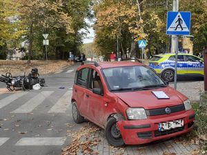 Skrzyżowanie na którym doszło do zdarzenia drogowego, samochód osobowy koloru czerwonego stoi na poboczu ma uszkodzony prawy przód, na środku skrzyżowania leży motocykl, a skrzyżowanie jest zabezpieczane przez parol policyjny