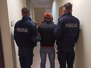 Mężczyzna w kapturze na głowie stoi tyłem do zdjęcia w korytarzu jednostki Policji. Po obu jego stronach stoją policjanci w umundurowaniu służbowym